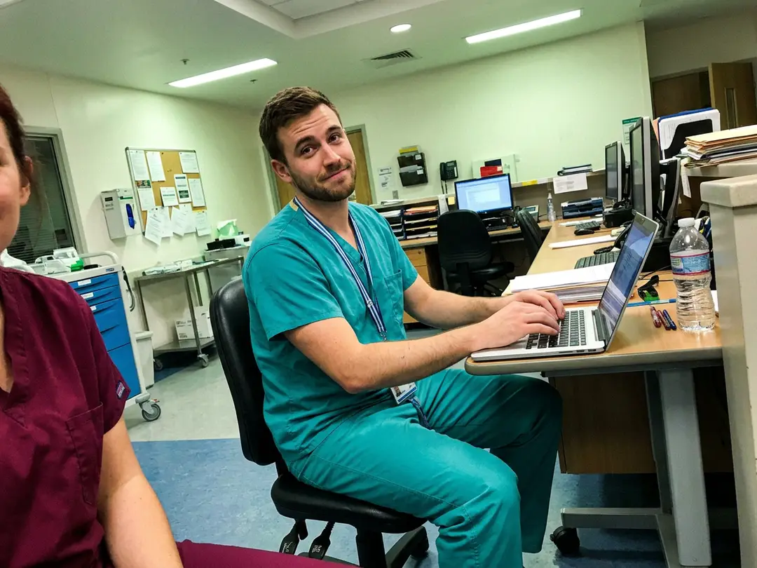 A nurse in teal scrubs at the nurses' station, working on a laptop