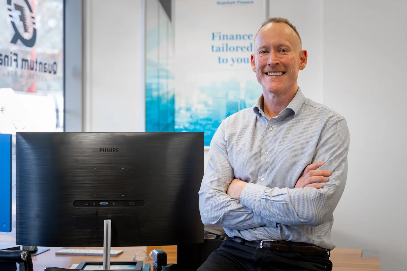 Gavin Harrigan standing with his arms folded beside a monitor at the West Leederville office, the Quantum Finance wall graphic behind him