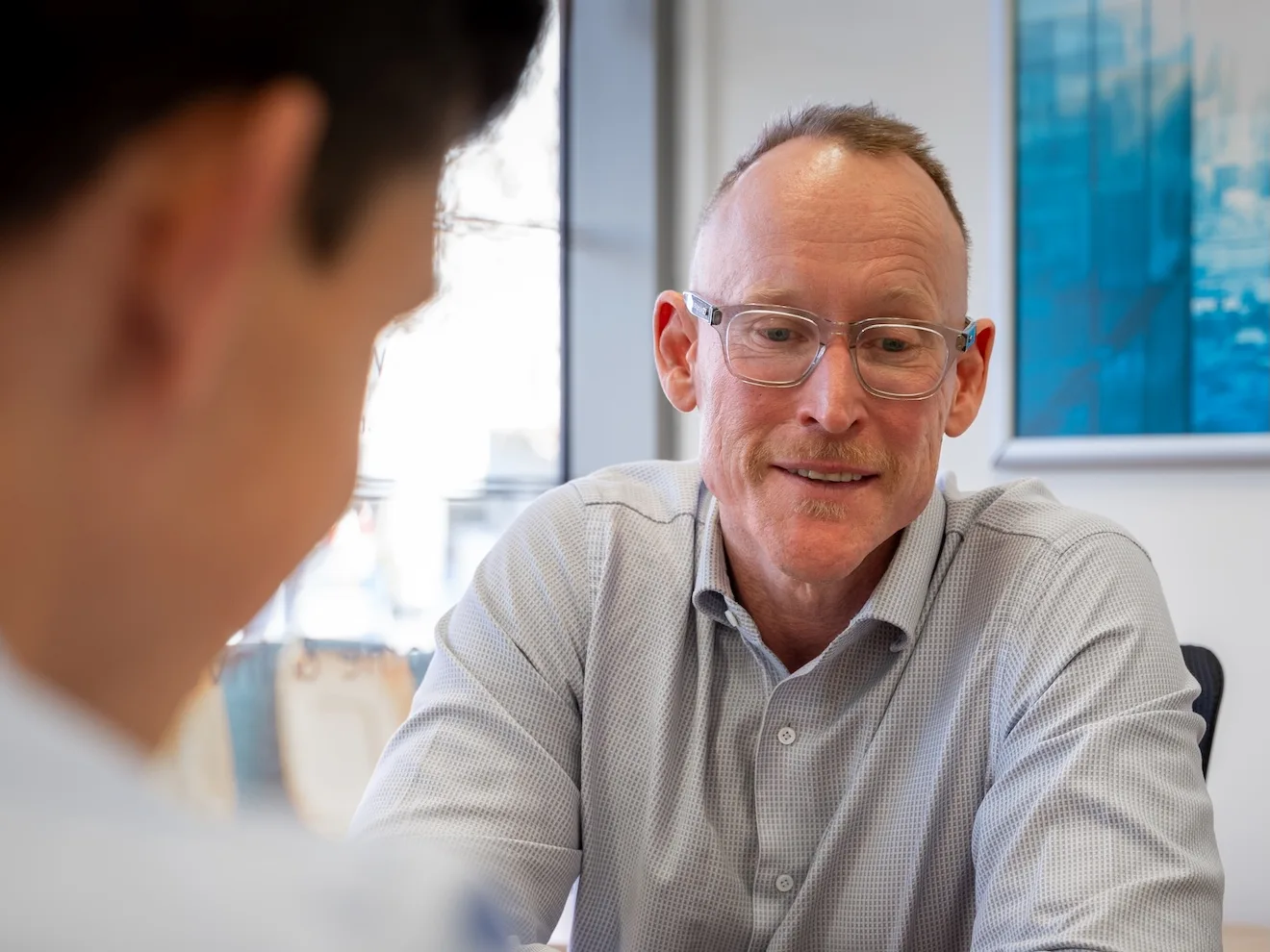 Gavin Harrigan in reading glasses listening to a client across the desk at the West Leederville office, seen past the client's shoulder