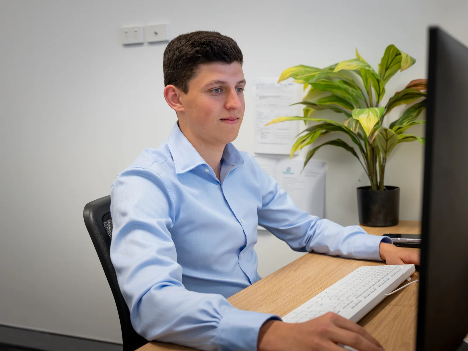 Xavier Prescott at his desk with both hands on the keyboard and his eyes on the monitor, pinned paperwork on the wall behind